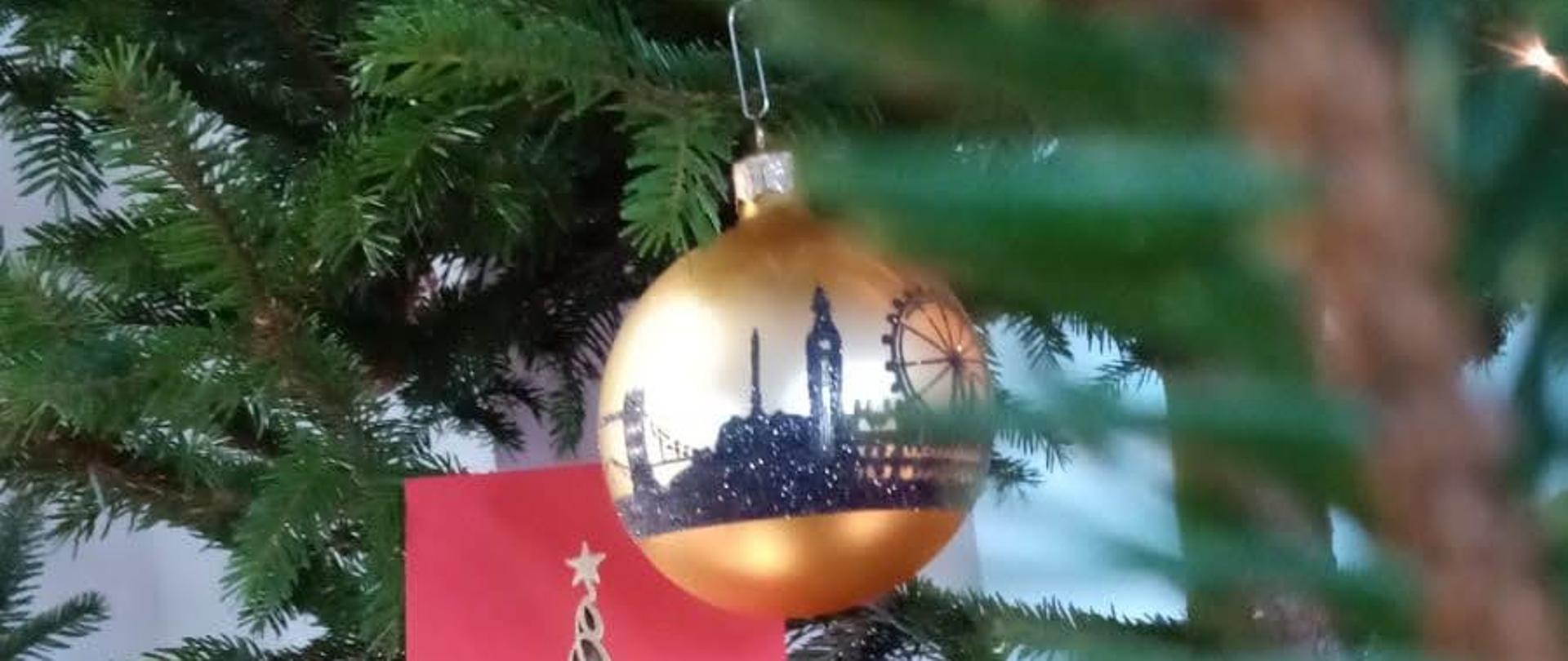Na zdjęciu znajduje się złota bombka z panoramą miasta zawieszoną na choince, wśród gałązek choinki umieszczona jest czerwona kartka ze złotą wstążką oraz grafiką złotej choinki, na gałązką umieszczone są świecące lampki choinkowe.