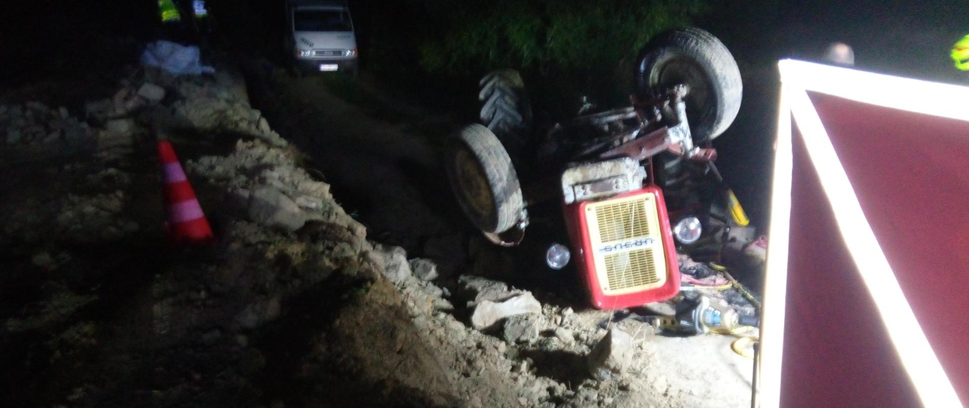 Zdjęcie przedstawia leżący na polnej drodze do góry kołami ciągnik rolniczy. Po prawej stronie widać fragment parawanu osłaniającego. 