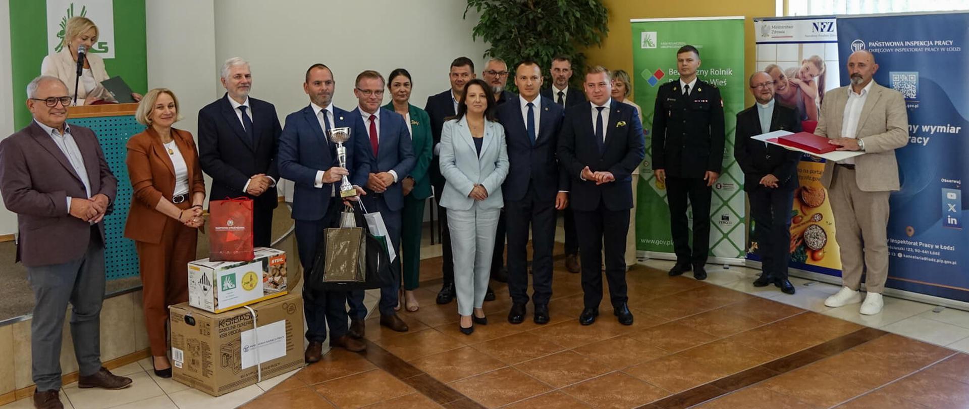 Uczestnicy i organizatorzy wydarzenia pozujący do pamiątkowego zdjęcia. Mężczyzna w garniturze trzyma puchar. Obok nich, na podłodze, ułożone są nagrody rzeczowe w kartonowych opakowaniach (m.in. agregat prądotwórczy).