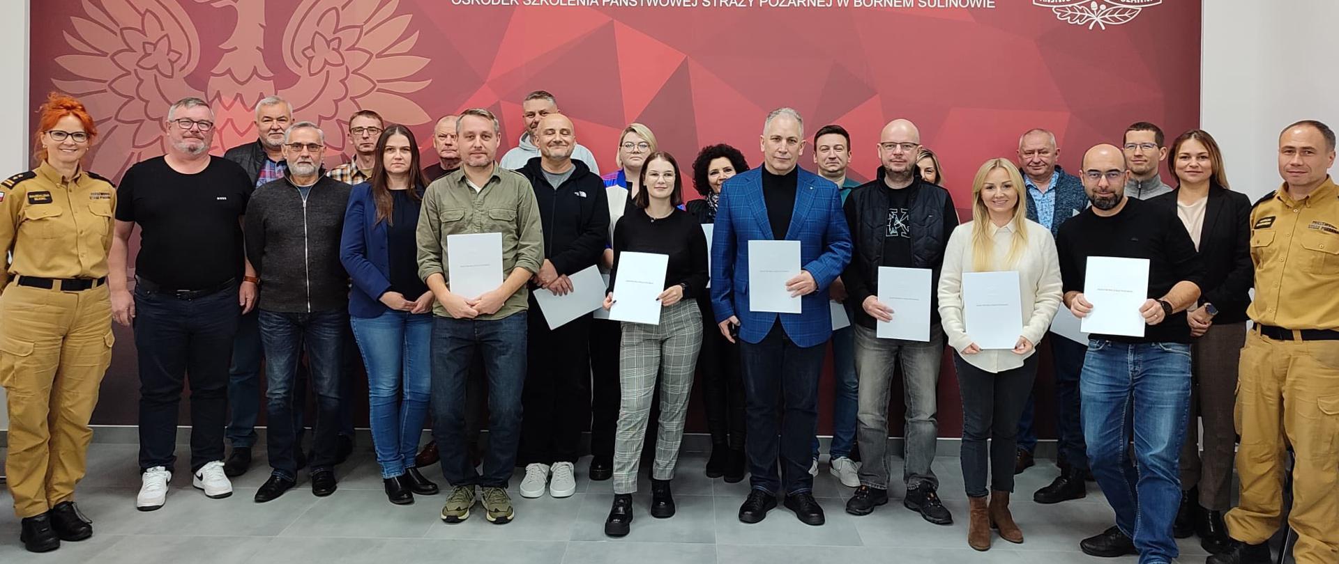 Na zdjęciu stoi duża grupa osób. Osoby stoją w dwóch rzędach i patrzą w stronę aparatu. Niektóre osoby trzymają w rękach białe teczki lub dokumenty. Po lewej i prawej stronie stoją dwie osoby w jasnych mundurach strażackich. W tle widać dużą, czerwoną tablicę z napisem: „Państwowa Straż Pożarna” oraz logo straży pożarnej.
