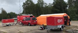 Samochody specjalne stoją obok namiotu przed namiotem stoi strażak obok przyczepka oraz agregat.