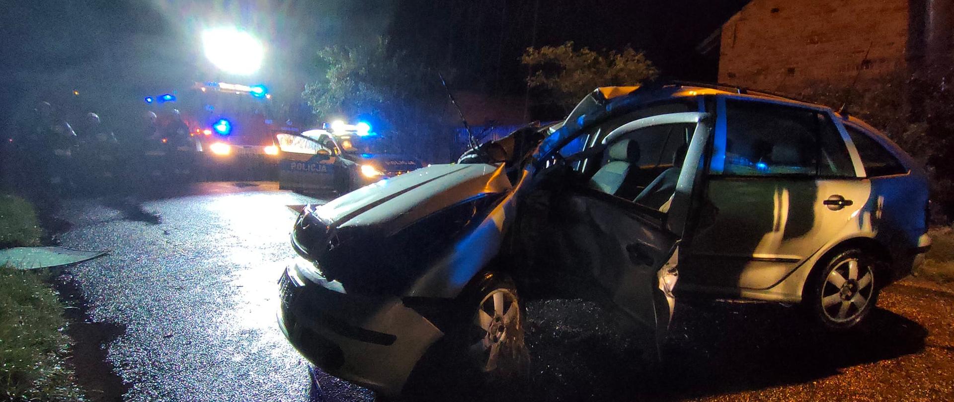 Widok samochodu z uszkodzonym bokiem. Pojazd wypadł z drogi i uderzył w drzewo. W tle pojazdy pożarnicze z włączonymi światłami błyskowymi. Obok pojazdów stoją strażacy.