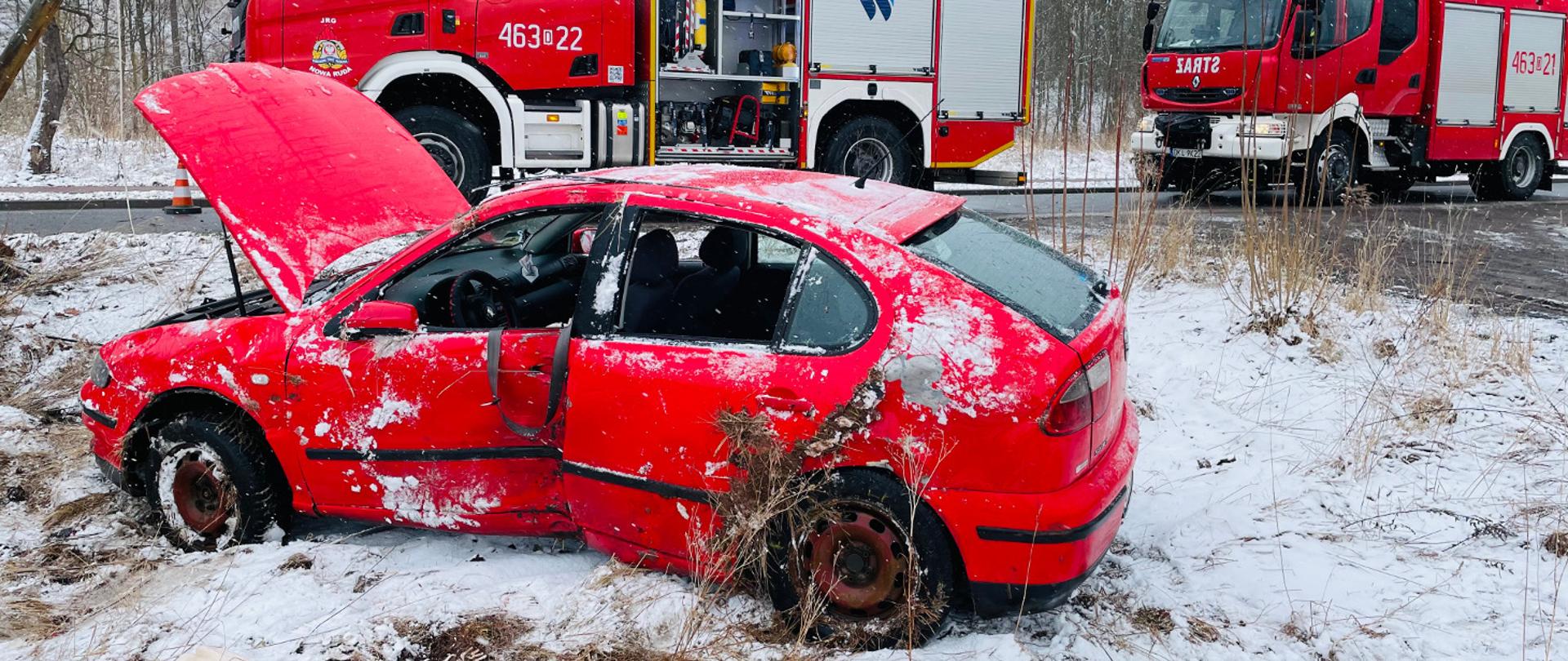 Na pierwszym planie uszkodzony samochód osobowy koloru czerwonego stojący na ośnieżonym poboczu. W tle dwa pojazdy Ochotniczych Straży Pożarnych.