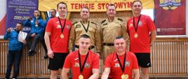 Pięciu zawodników w czerwonych koszulkach z medalami pozuje z dwoma funkcjonariuszami PSP w beżowych mundurach. Z przodu stoją puchar i dyplom. W tle widoczny baner zawodów i roll-upy partnerów.
Tekst na banerze: „XXV Mistrzostwa Województwa Podkarpackiego Strażaków PSP w tenisie stołowym”.
