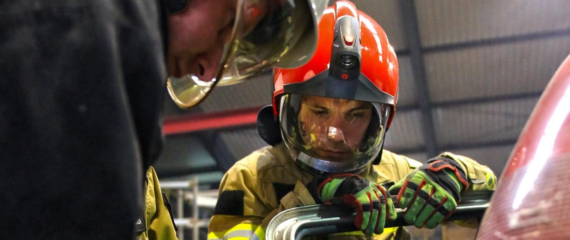 Na zdjęciu widać strażaka w umundurowaniu bojowym koloru musztardowego. Strażak ma na głowie hełm koloru czerwonego. Strażak to instruktor ratownictwa technicznego obok niego znajduje się strażak ochotniczej straży pożarnej, trzymający urządzenie. Zdjęcie wykonane w hali. 