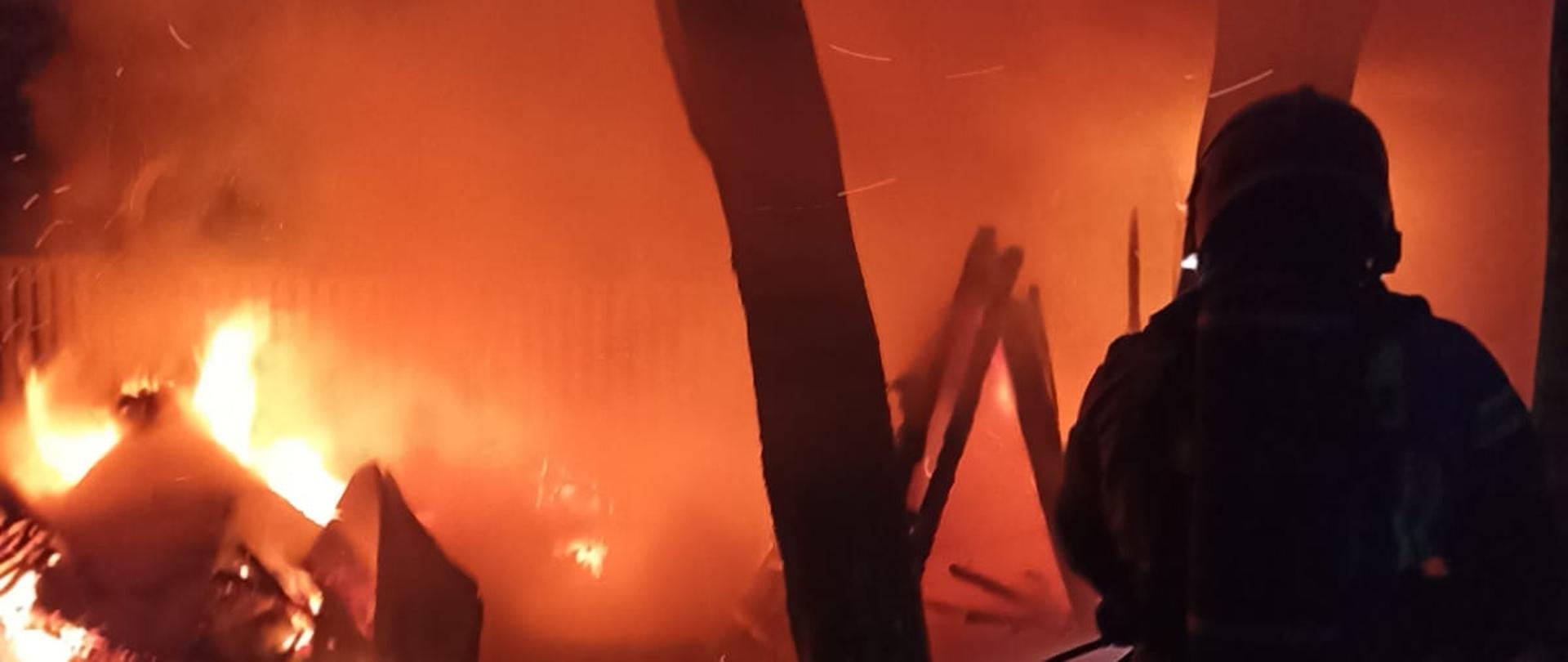 Na posesji w nocy pali się niewielki budynek gospodarczy. Obok betonowe ogrodzenie i drzewa. Z boku sylwetka strażaka.