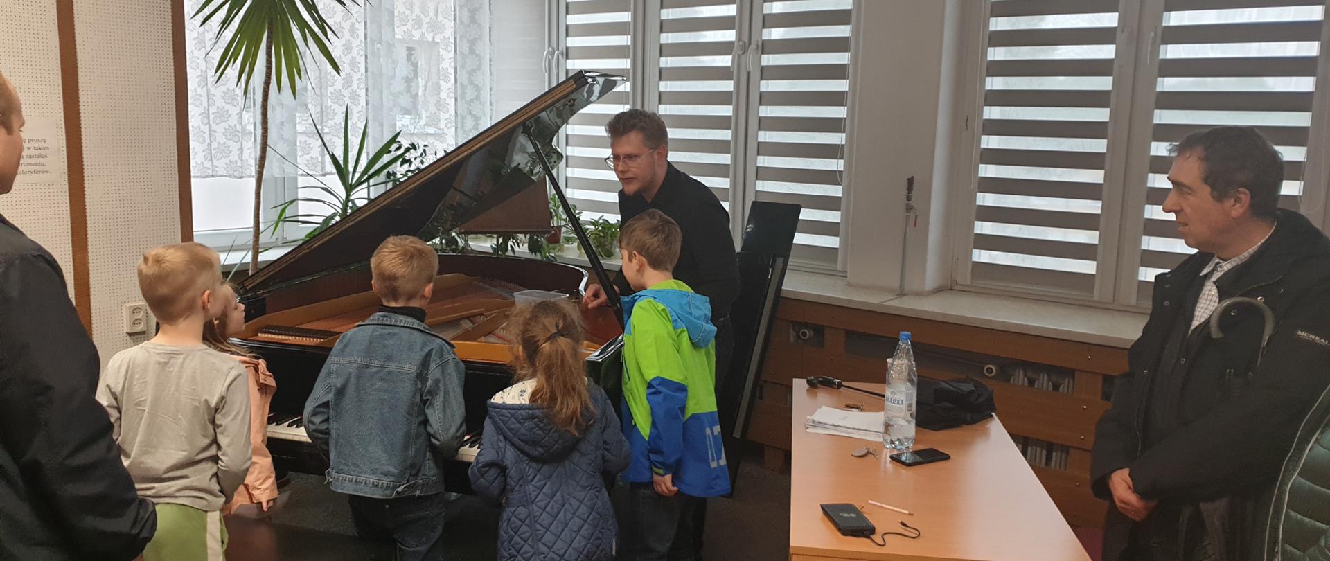 Pięcioro dzieci stoi przy fortepianie i ogląda jego wnętrze - struny, obok stoi mężczyzna i mówi do dzieci. Po bokach stoi dwoje rodziców.