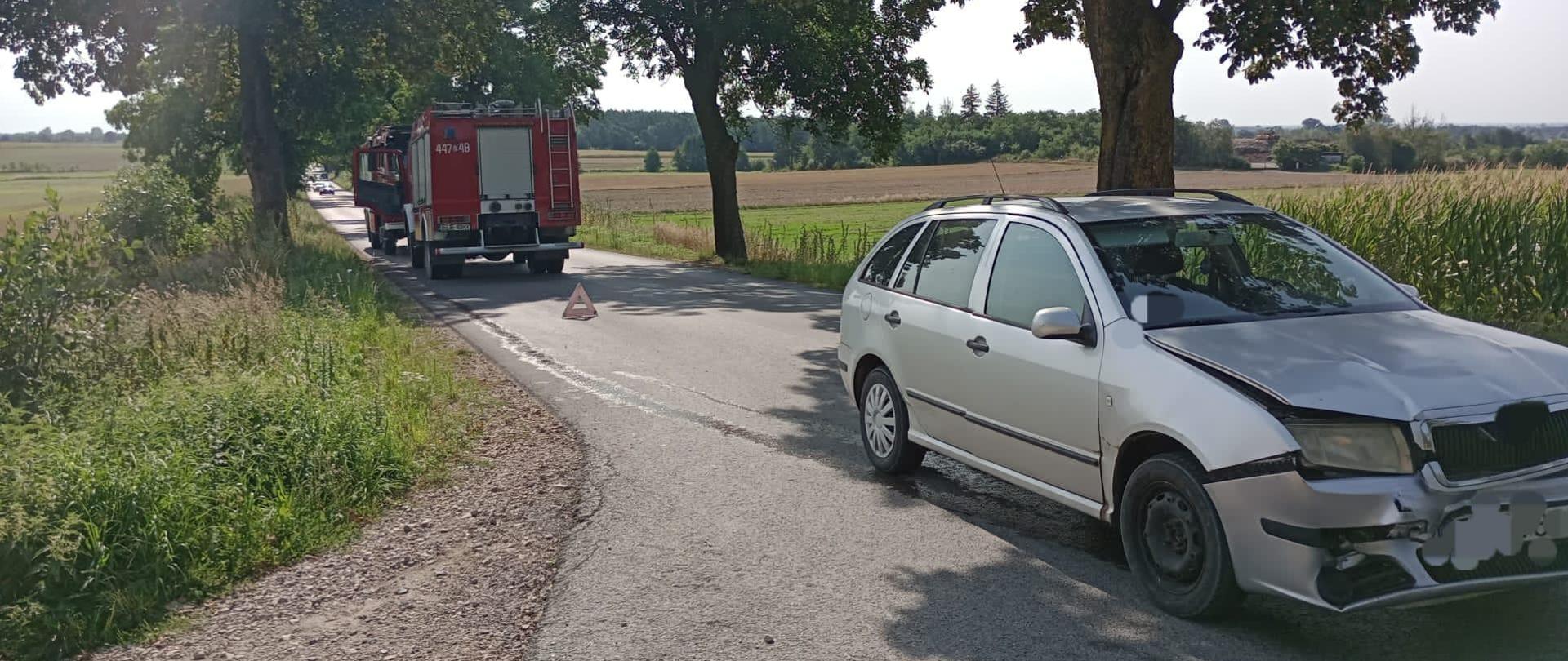 Na zdjęciu widać srebrny samochód z uszkodzonym przodem, który stoi na asfaltowej drodze. Z tyłu widać dwa samochody strażackie oraz ustawiony trójkąt ostrzegawczy. Dookoła widać pola, drzewa i krzaki. Jest słonecznie.