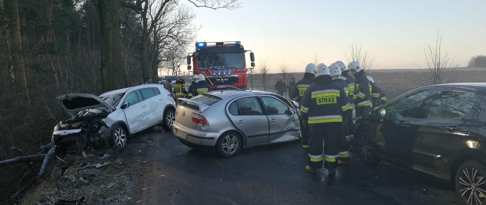 Zdjęcie przedstawia kolizję drogową z udziałem dwóch samochodów osobowych na drodze. Na zdjęciu widać grupę ratowników podczas działań. Na dalszym planie widoczny wóz ratowniczo- gaśniczy przed którym stoi grupa strażaków. Zdjęcie wykonane w porze dziennej na drodze asfaltowej.