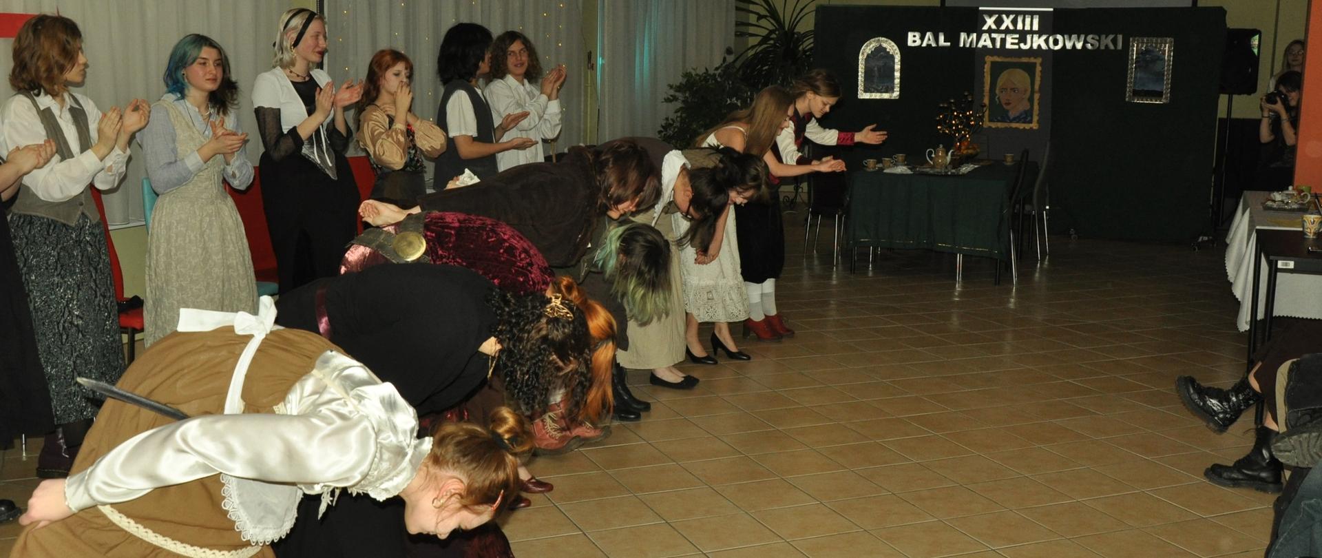 Grupa kilkunastu osób przebranych w stroje sceniczne kłania się publiczności. W tle scenografia z napisem XXIII Bal Matejkowski