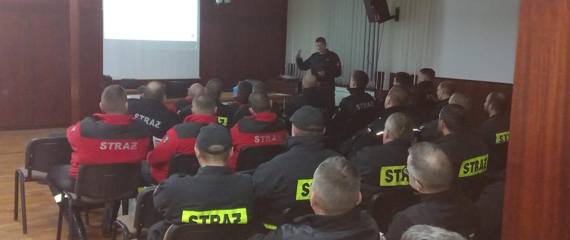 Strażaka który przedstawia za pomocą rzutnika prezentację dla druhów OSP. Strażak stoi i trzyma w ręku pilot Druhowie siedzą. Na stoliku położony jest laptop połączony z rzutnikiem za pomocą którego wyświetlana jest prezentacja.