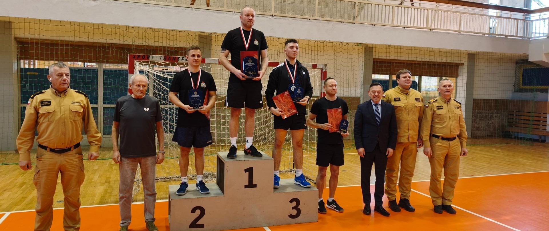 Ceremonia wręczenia nagród w hali sportowej. Trzech mężczyzn stoi na podium oznaczonym miejscami 1, 2 i 3, trzymając puchary i dyplomy. Wokół nich stoją inni uczestnicy oraz trzech funkcjonariuszy w umundurowaniu służbowym. W tle widoczna jest siatka bramki oraz dekoracyjna ściana z motywami sportowymi.