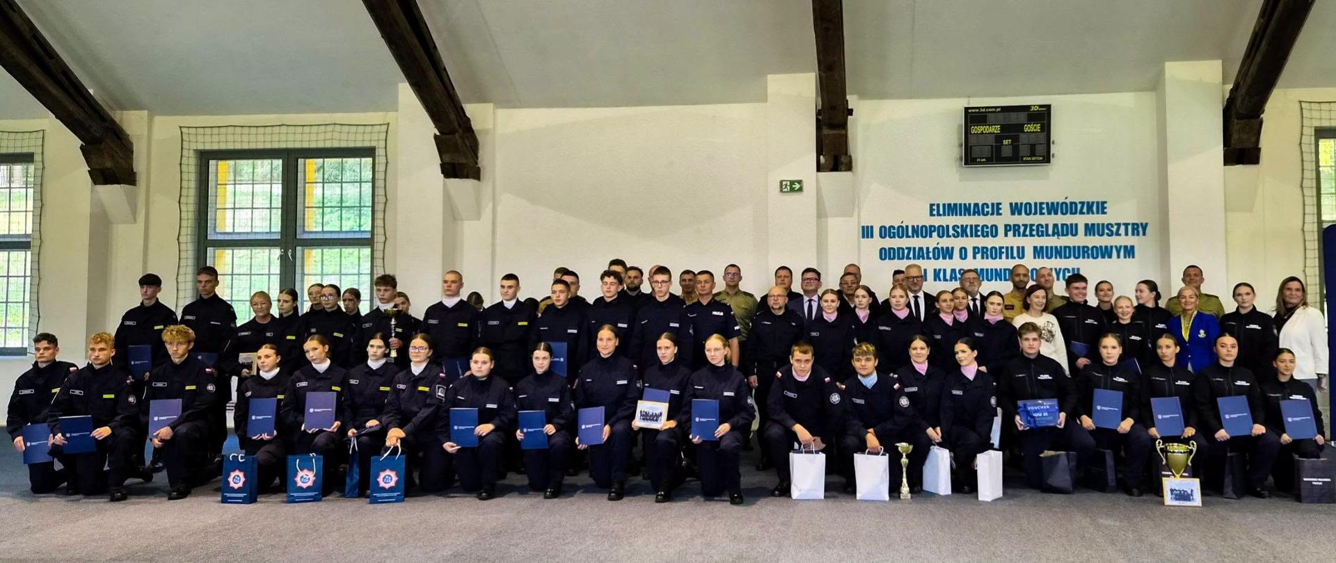 Zdjęcie grupowe przedstawiające młodych funkcjonariuszy w mundurach. W ostatnim rzędzie stoją przedstawiciele władz i organizatorzy wydarzenia, wśród nich wojewoda warmińsko-mazurski.
