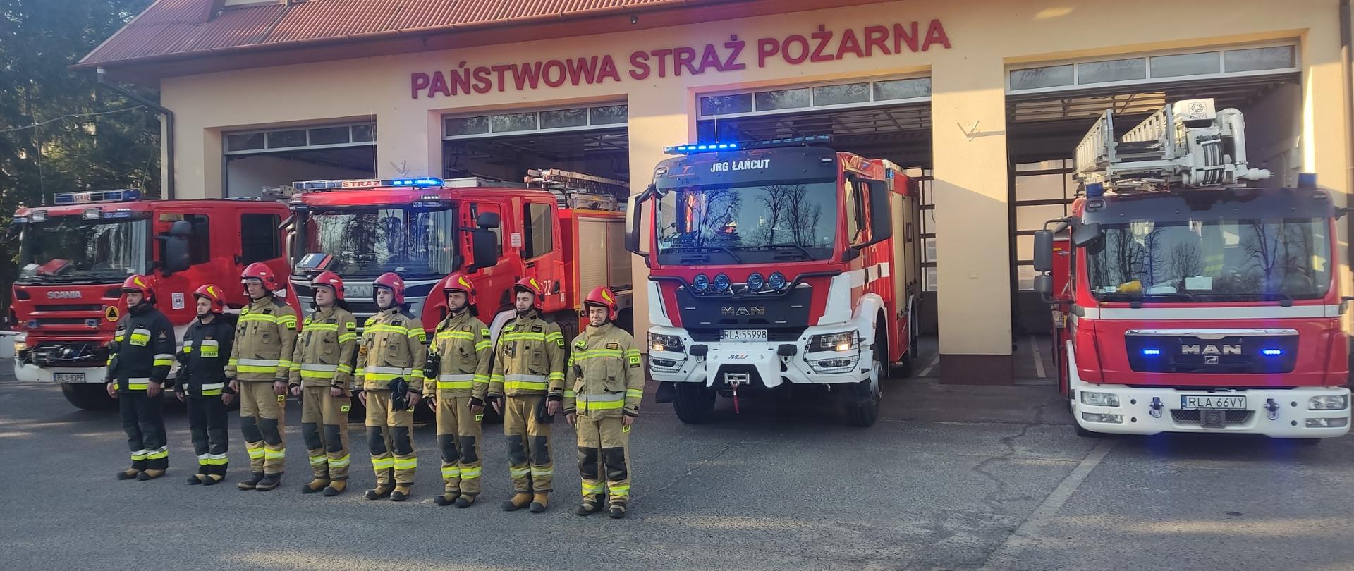 Zdjęcie przedstawia plac zewnętrzny komendy na którym ustawione są cztery samochody Komendy Powiatowej. Na pierwszym planie stoją strażacy Komendy Powiatowej Państwowej Straży Pożarnej w Łańcucie.