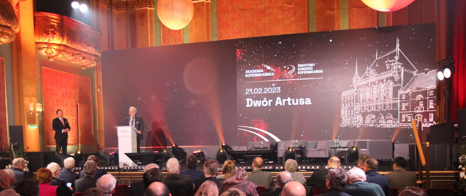 Widok na salę z tyłu, na krzesłach siedzą ludzie, patrzą na wprost. Przy pulpicie stoi mężczyzna i przemawia. 