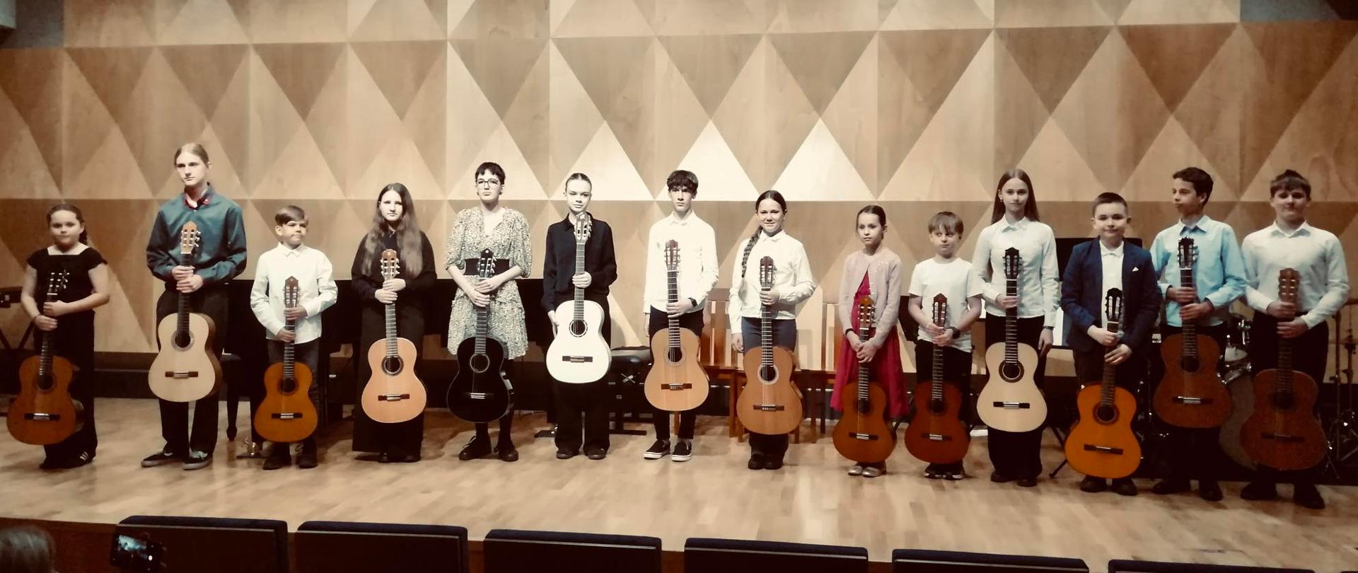 Grupa uczniów z gitarami w rękach na scenie sali koncertowej