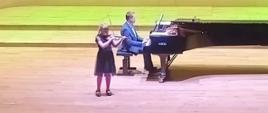 Dziewczynka gra na skrzypcach, za Nią mężczyzna gra na fortepianie.