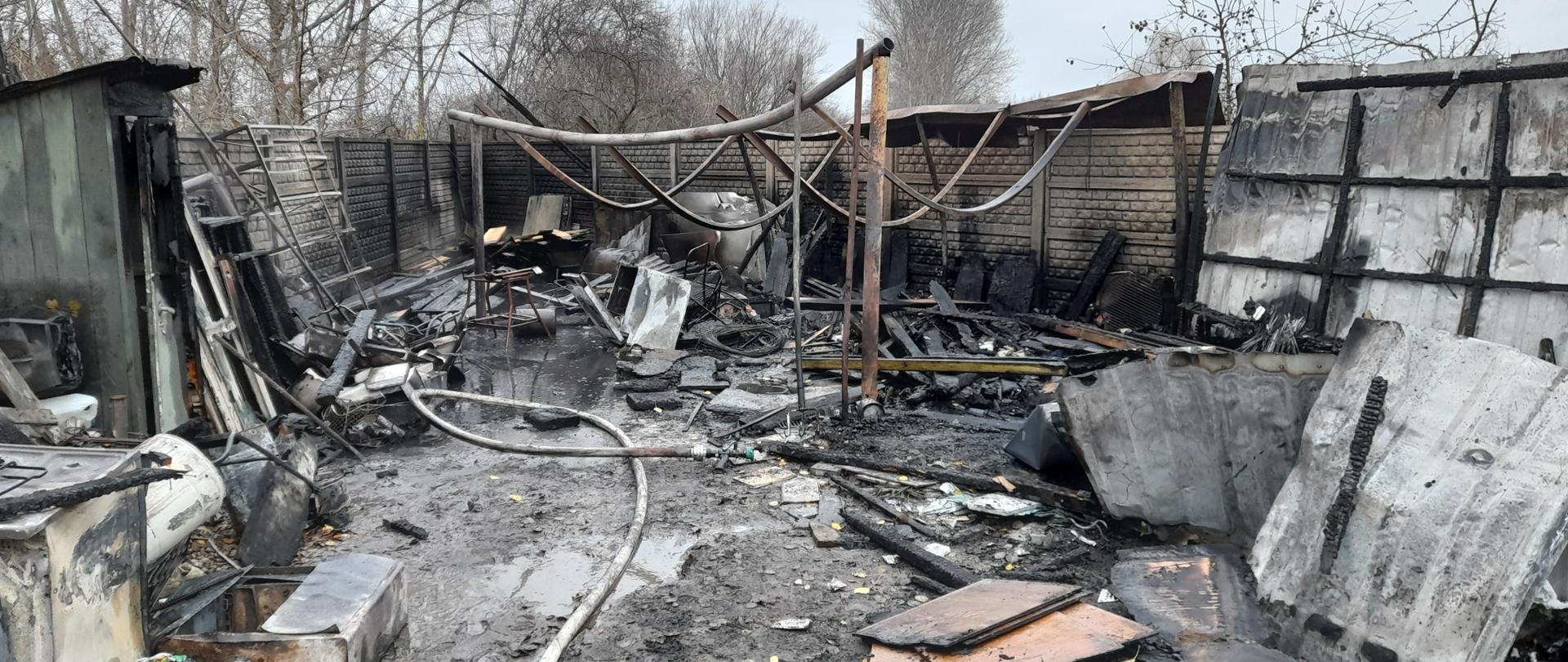 Zdjęcie przedstawia całkowicie spalony garaż wraz z wyposażeniem. Na zdjęciu widać ogrodzenie z płyt betonowych oraz pozostałości konstrukcji stalowej garażu 