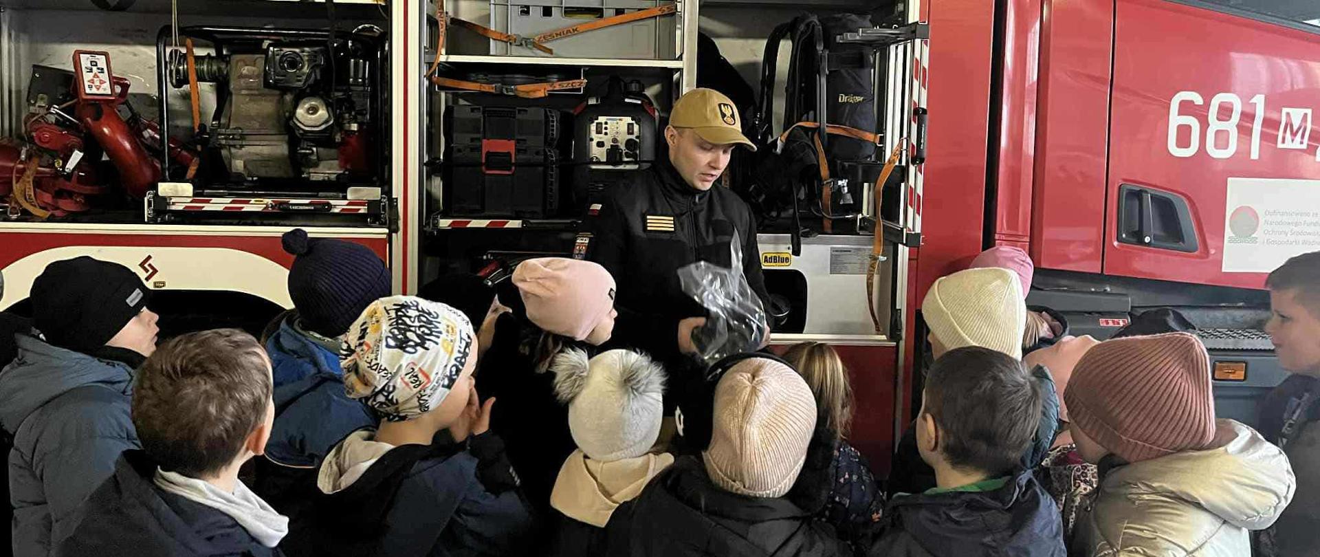 „Strażak pokazuje dzieciom wyposażenie wozu strażackiego, otwierając boczną skrytkę pełną sprzętu ratowniczego. Grupa dzieci w zimowych kurtkach stoi blisko i uważnie słucha.”