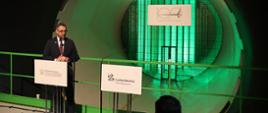 Za mównicą z napisem Łukasiewicz stoi minister Kulasek w czarnym garniturze, mówi do mikrofonu, za nim oświetlony na zielono osłonięty siatką wlot tunelu.