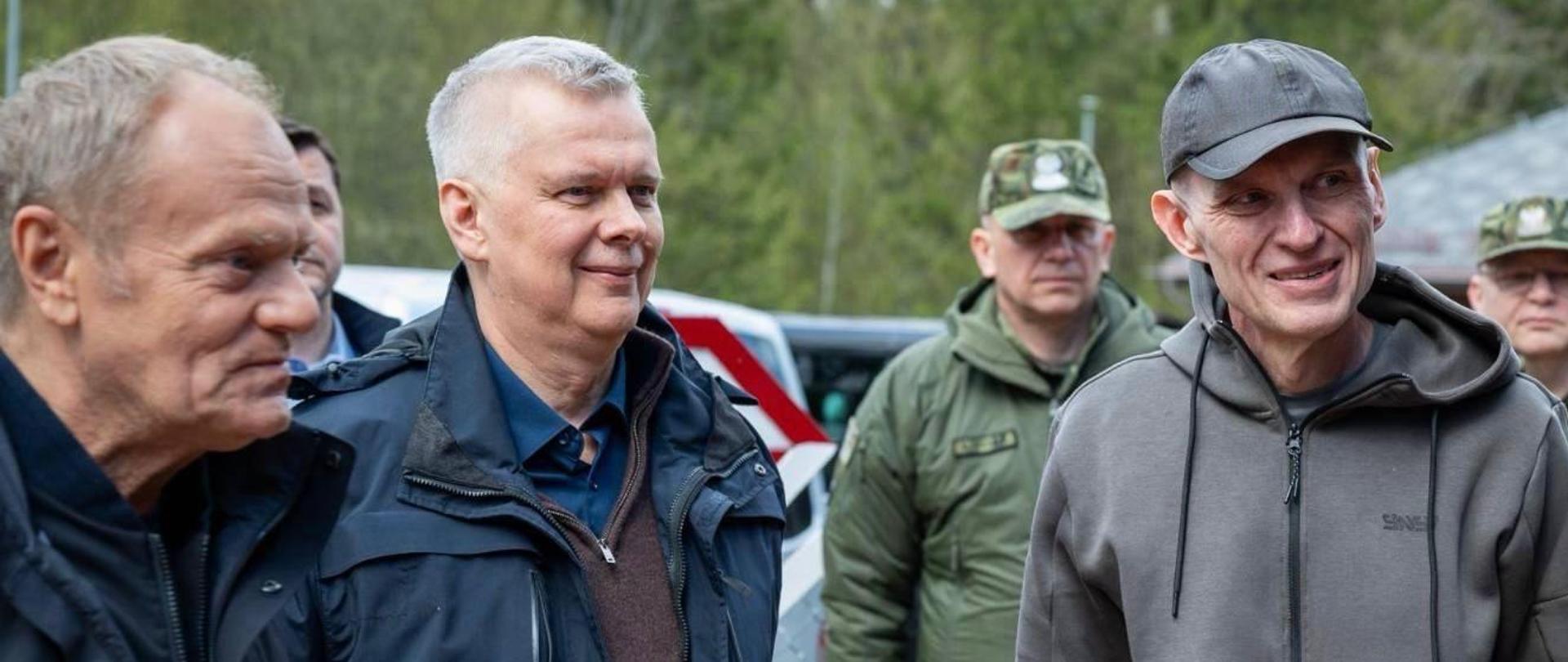 In the foreground, three people - from the right Andrzej Poczobut, Minister Tomasz Siemoniak, and Prime Minister Donald Tusk. Behind Andrzej Poczobut stand two Border Guard officers. In the background, cars and trees can be seen.