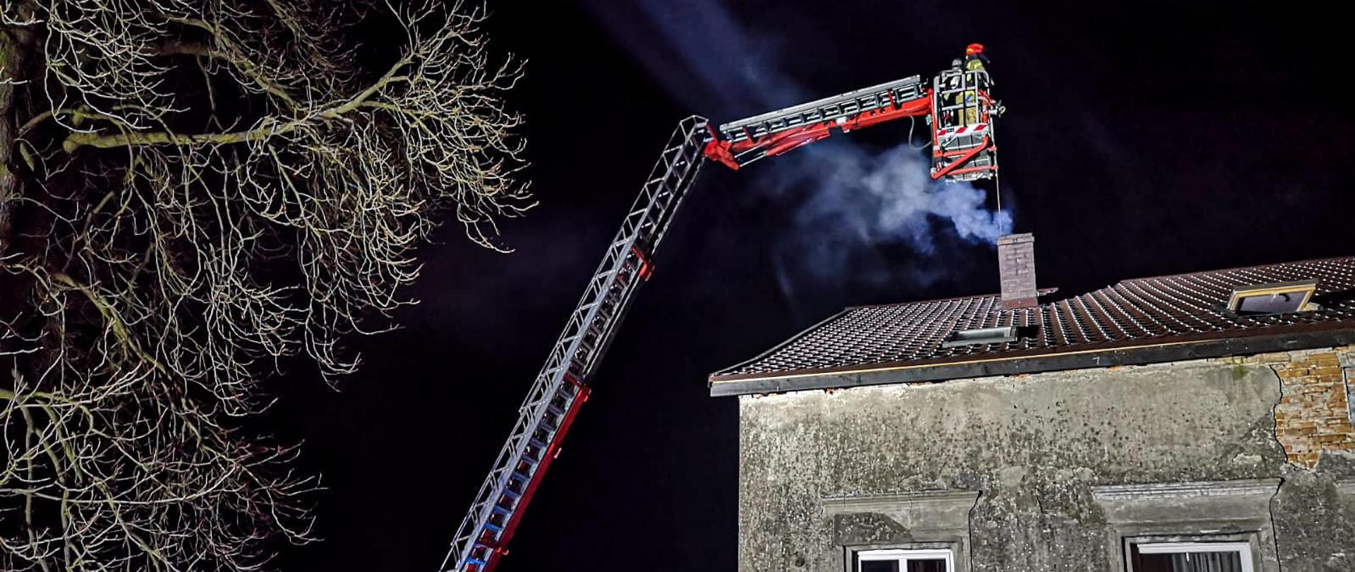 Na zdjęciu wykonanym w porze nocnej na pierwszym planie znajduje się budynek z dwoma oknami. Po lewej stronie domu rozstawiony jest strażacki podnośnik, w koszu którego znajdują się strażacy. Pod koszem podnośnika znajduje się komin z którego wydobywa się dym. Po lewej stronie znajduje się duże drzewo.