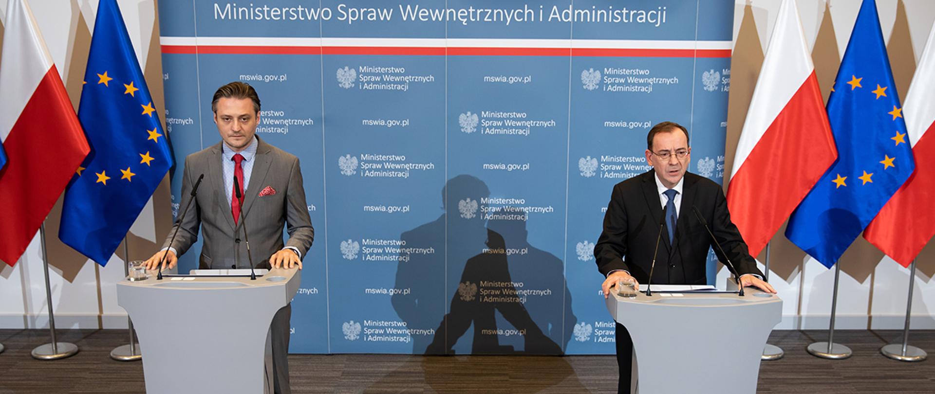 Konferencja prasowa ministra Mariusza Kamińskiego i wiceministra Bartosza Grodeckiego. Widać obu ministrów na tle ścianki MSWiA i flag Polski i Uni Europejskiej przy mównicach. Minister Mariusz Kamiński zabiera głos.