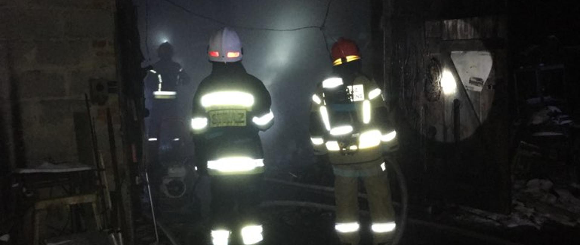 1. Zdjęcie przedstawia budynek murowany garażu z otwartymi drzwiami oraz trzech strażaków prowadzących działania gaśnicze wewnątrz niego. Jeden ze strażaków ubrany jest w sprzęt ochrony układu oddechowego trzymający w dłoniach odcinek linii gaśniczej. Wnętrze garażu jest zadymione. Na zewnątrz liczne elementy metalowe oparte o ściany garażu.