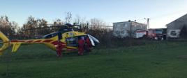 Zdjęcie przedstawia, stojący na zielonym polu, żółto-czerwony śmigłowiec, obok którego chodzą członkowie załogi. W tle zabudowania i srebrno-czerwony strażacki samochód ciężarowy.