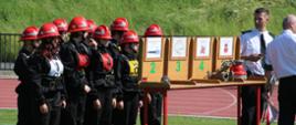 po lewej stronie dziewięciu uczestników zawodów ubranych w czarne mundury i czerwone hełmy, po prawej stronie dwóch mężczyzn - organizatorów zawodów, ubranych w mundury, na środku stół z ponumerowanymi przegrodami, w przegrodach przedmioty będące w użyciu podczas pracy straży pożarnej