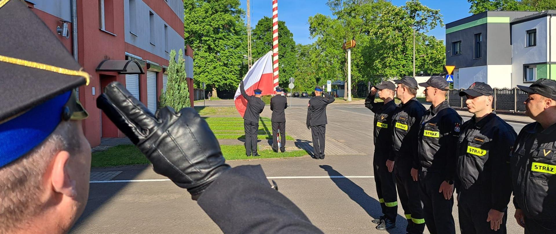 Na zdjęciu widoczna jest uroczystość podniesienia flagi państwowej przed budynkiem Komendy Powiatowej Państwowej Straży Pożarnej . Po lewej stronie budynek z czerwonej cegły. Przy maszcie strażacy w galowych mundurach zawieszają flagę. z lewej strony strażacy w czarnych mundurach stoją w szeregu 
