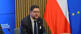 Wiceminister Szeptycki siedzi za stołem, mówi do małego mikrofonu, za nim pod ścianą flagi Polski i UE.