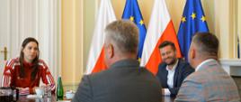 Minister Sportu i Turystyki Kamil Bortniczuk siedzi przy owalnym stole wraz z Martyną Swatowską-Wenglarczyk, polską szpadzistką, którą zdobyła drużynowe złoto na mistrzostwach świata w Mediolanie. Obok ministra siedzącego na szczycie siedzi również prezes Polskiego Komitetu Olimpijskiego Radosław Piesiewicz i przedstawiciele władz Polskiego Związku Szermierczego. W tle stoją flagi Polski i Unii Europejskiej.