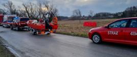 Zdjęcie przedstawia zaparkowane na poboczu drogi samochody ratownicze, tj. samochód operacyjny Państwowej Straży Pożarnej, lekki samochód rozpoznawczo-ratowniczy z przyczepką, na której znajduje się łódź oraz karetkę pogotowia. Na drugim planie w głębi łąki widnieje rozłożony parawan ochronny w czerwonym kolorze.