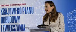 Minister funduszy i polityki regionalnej Katarzyna Pełczyńska-Nałęcz stoi przy pulpicie i przemawia do mikrofonu, za jej plecami grafika z napisem: "Spotkanie roczne w sprawie Krajowego Planu Odbudowy i Zwiększenia Odporności" na tle mapy Polski 