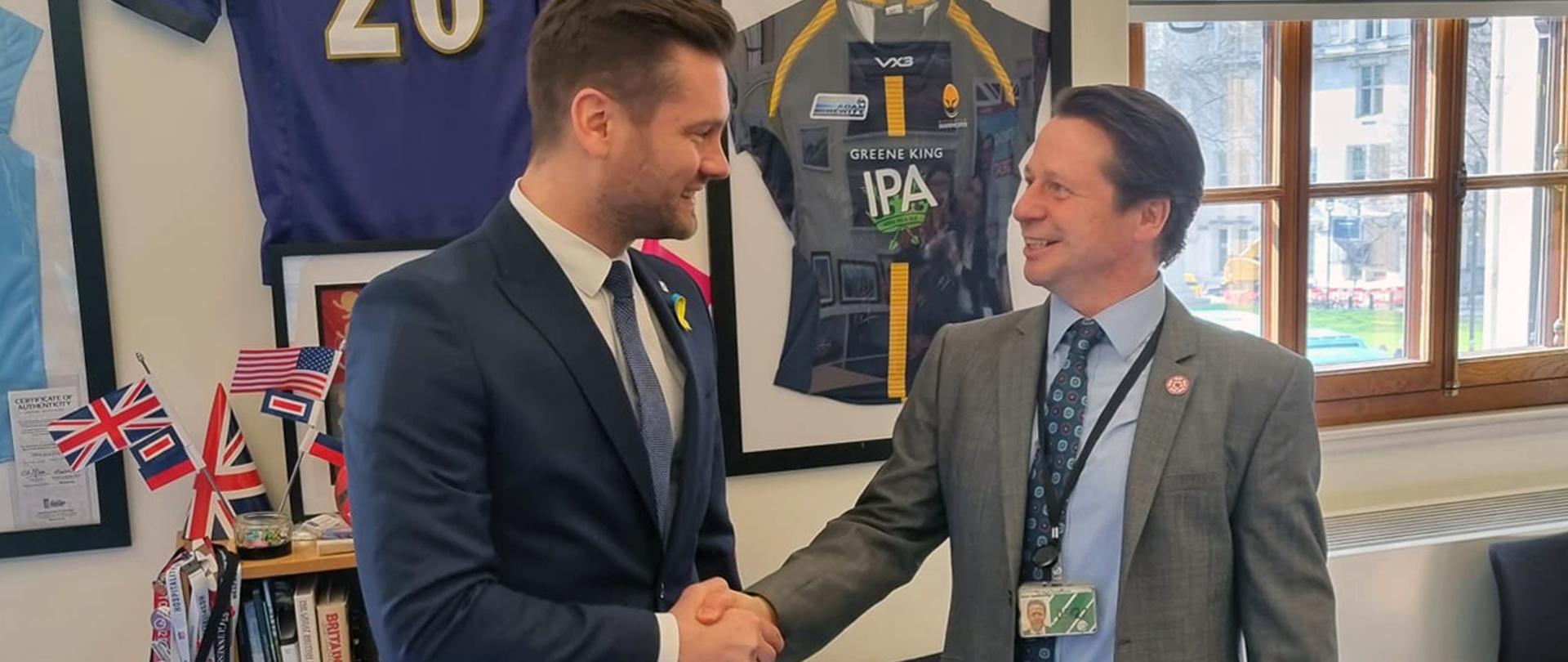 Ministrowie sportu Polski i Wielkiej Brytanii omówili w Londynie sprawę dalszych sankcji dla Rosji w obszarze sportu. Na zdjęciu minister sportu i turystyki Kamil Bortniczuk oraz minister Wielkiej Brytanii do spraw sportu Nigel Huddlestone podają sobie ręce. Na ścianach widoczne koszulki sportowe, w tle ścianka z książkami i flagami Wielkiej Brytanii.