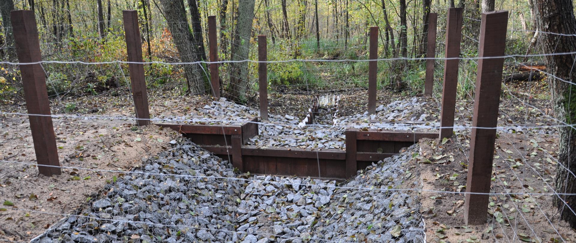 Drewniano-kamienna konstrukcja ustawiona pośród drzew. Dno konstrukcji wypełnione kamieniami. W tle niewielki rów z wodą.