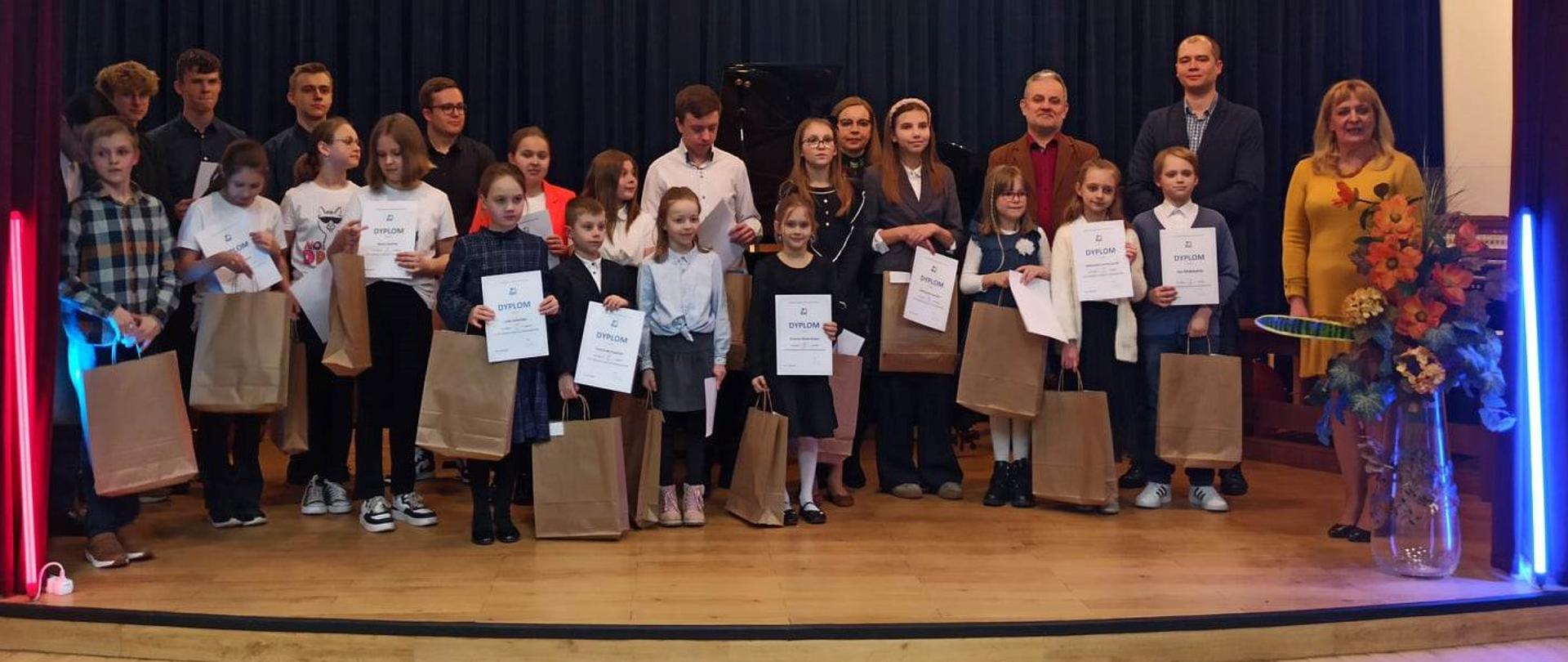 Zdjęcie przedstawia uczniów stojących na scenie z prezentami oraz uniesionymi dyplomami. Po prawej stronie jest trzech nauczycieli. 