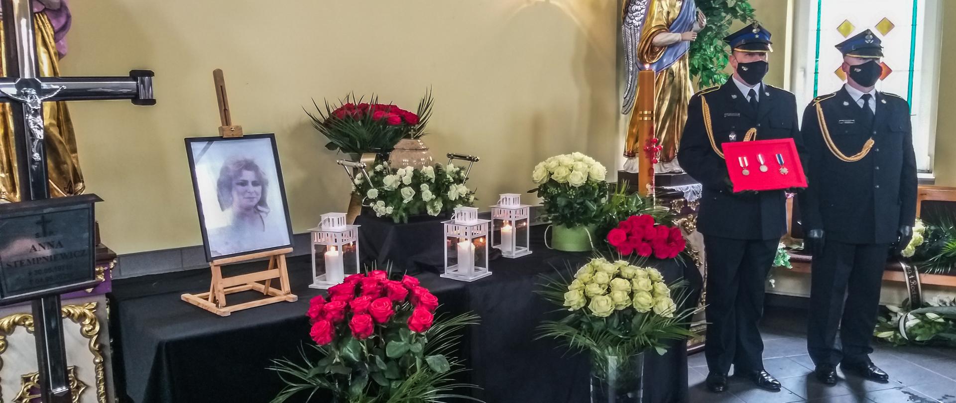 Zdjęcie wykonane w trakcie uroczystości pogrzebowych. Urna z prochami zmarłej otoczona lampionami i wiązankami z kwiatów. Po lewej stronie krzyż z tabliczką. Obok urny zdjęcie zmarłej. Po prawej stronie funkcjonariusz trzymający odznaczenia zmarłej. Obok drugi funkcjonariusz asystuje mu. Obaj ubrani w mundury galowe.