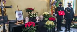 Zdjęcie wykonane w trakcie uroczystości pogrzebowych. Urna z prochami zmarłej otoczona lampionami i wiązankami z kwiatów. Po lewej stronie krzyż z tabliczką. Obok urny zdjęcie zmarłej. Po prawej stronie funkcjonariusz trzymający odznaczenia zmarłej. Obok drugi funkcjonariusz asystuje mu. Obaj ubrani w mundury galowe.