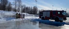 Dwóch funkcjonariuszy PSP w ubraniach specjalnych podają jeden prąd wody z samochodu gaśniczego na boisko sportowe, na którym jest utworzone sztuczne lodowisko. 
