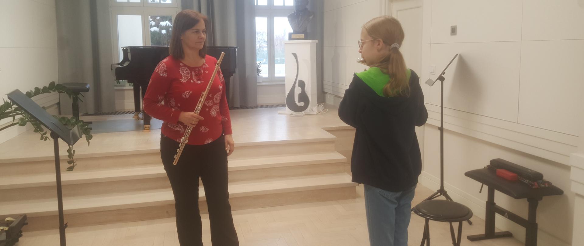 Na pierwszym planie z lewej osoba prowadząca warsztaty fletowe ubrana w czerwoną bluzkę i trzymająca flet, z prawej uczennica w granatowej bluzie z kapturem grająca na flecie. W tle po lewej pulpit na nuty, a dalej fortepian, z prawej postument z popiersiem patrona szkoły. Z tyłu odsłonięte okna z kotarami (zasłonami w szarym kolorze). 