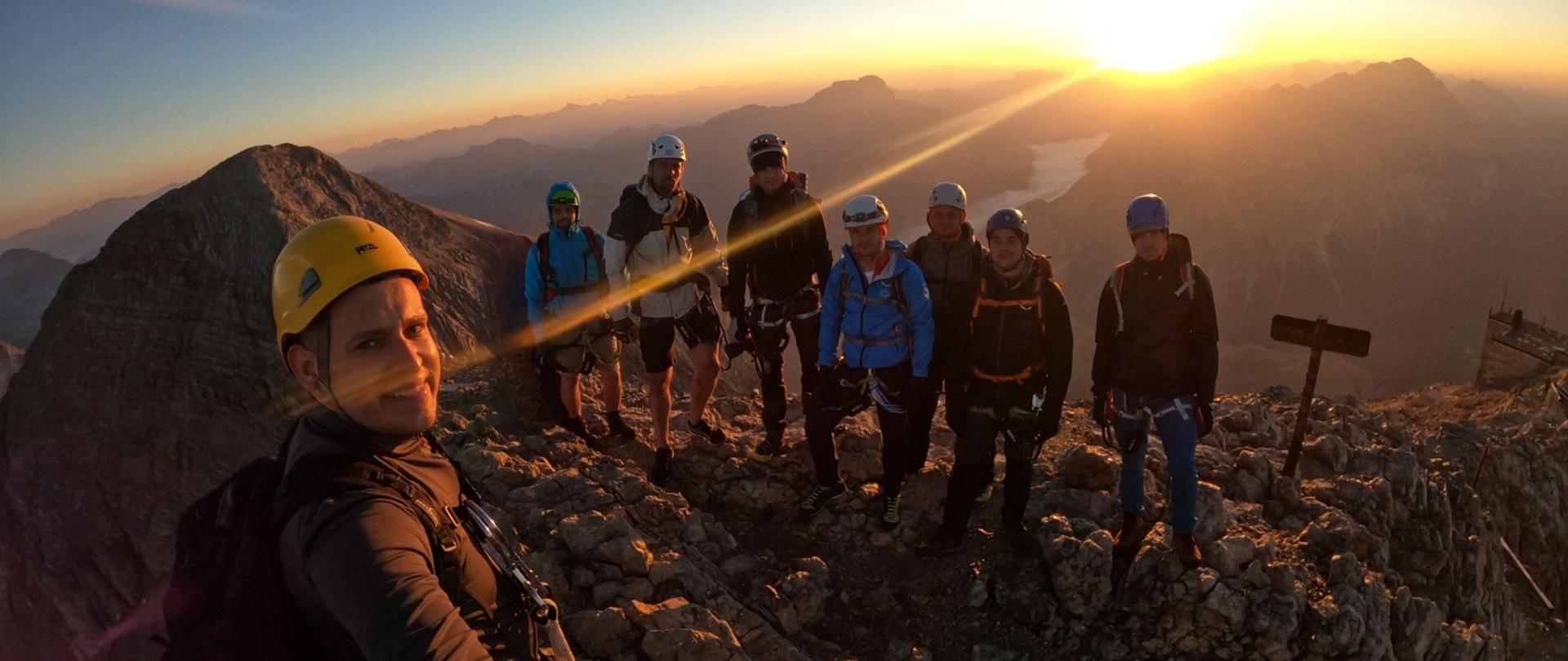 Grupa ośmiu alpinistów na szczycie górskim o wschodzie słońca, podczas wyprawy wysokogórskiej. W tle rozciąga się panorama gór. W lewym górnym rogu znajduje się logo fundacji Dorastaj z Nami, a w prawym – znak Państwowej Straży Pożarnej z informacją o patronacie honorowym Komendanta Głównego PSP.