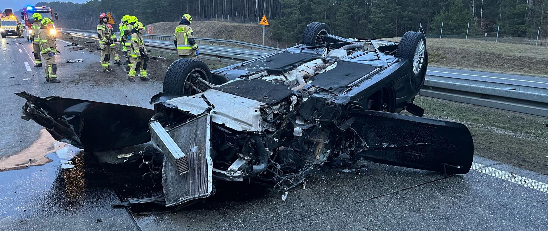 Samochód osobowy przewrócony na dach, zniszczony przód, otwarte drzwi, w tle strażacy