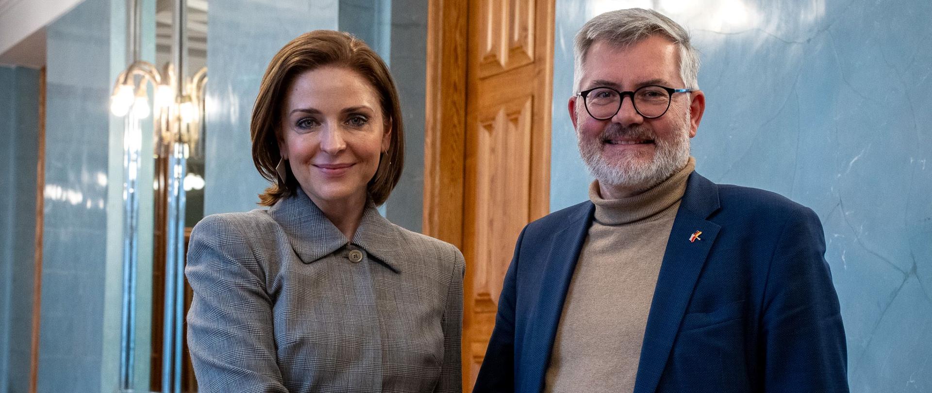 Minister Joanna Mucha stoi obok mężczyzny w garniturze i podają sobie ręce.