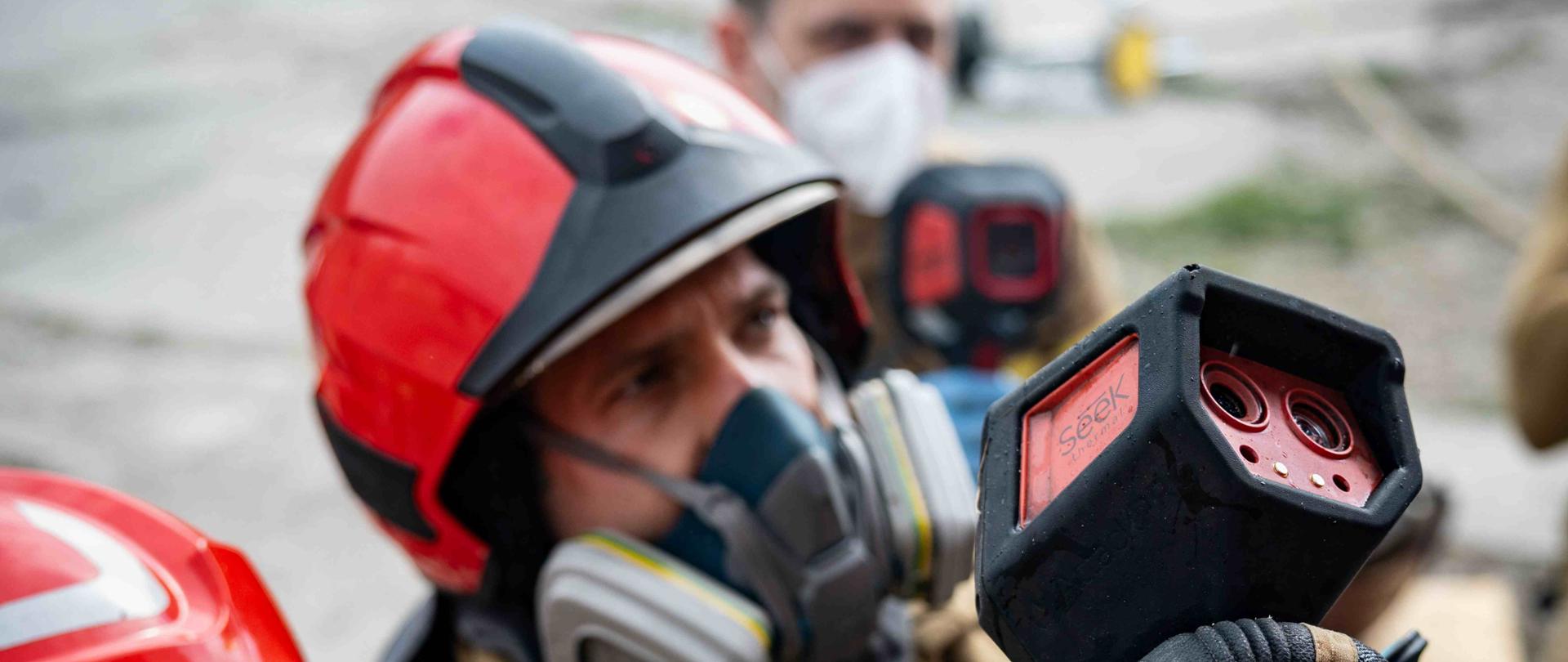 Strażacy w ubraniach specjalnych i maskach ochronnych pracują w terenie, obserwując wskazania kamery termowizyjnej. Jeden z nich trzyma urządzenie, a pozostali uważnie przyglądają się jego działaniu.