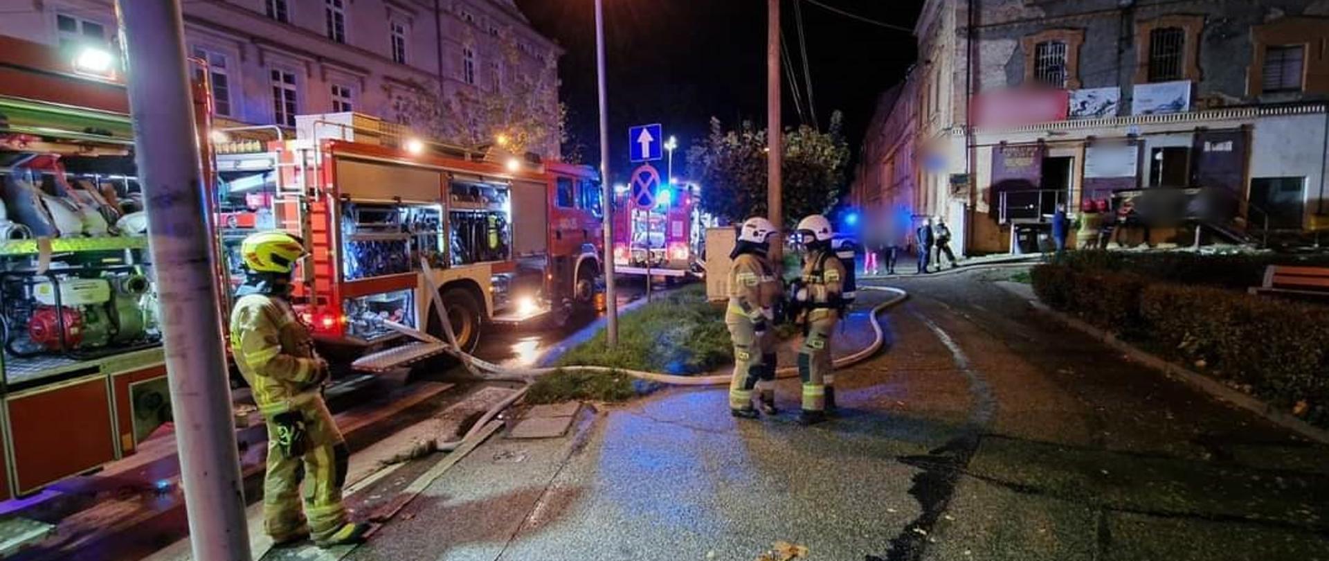 Obraz przedstawia strażaków w ubraniach specjalnych prowadzących działania gaśnicze przy ul. Kolejowej. Obok pojazdy pożarnicze z włączonym oświetleniem alarmowym, w tle budynki mieszkalne. Pora nocna.