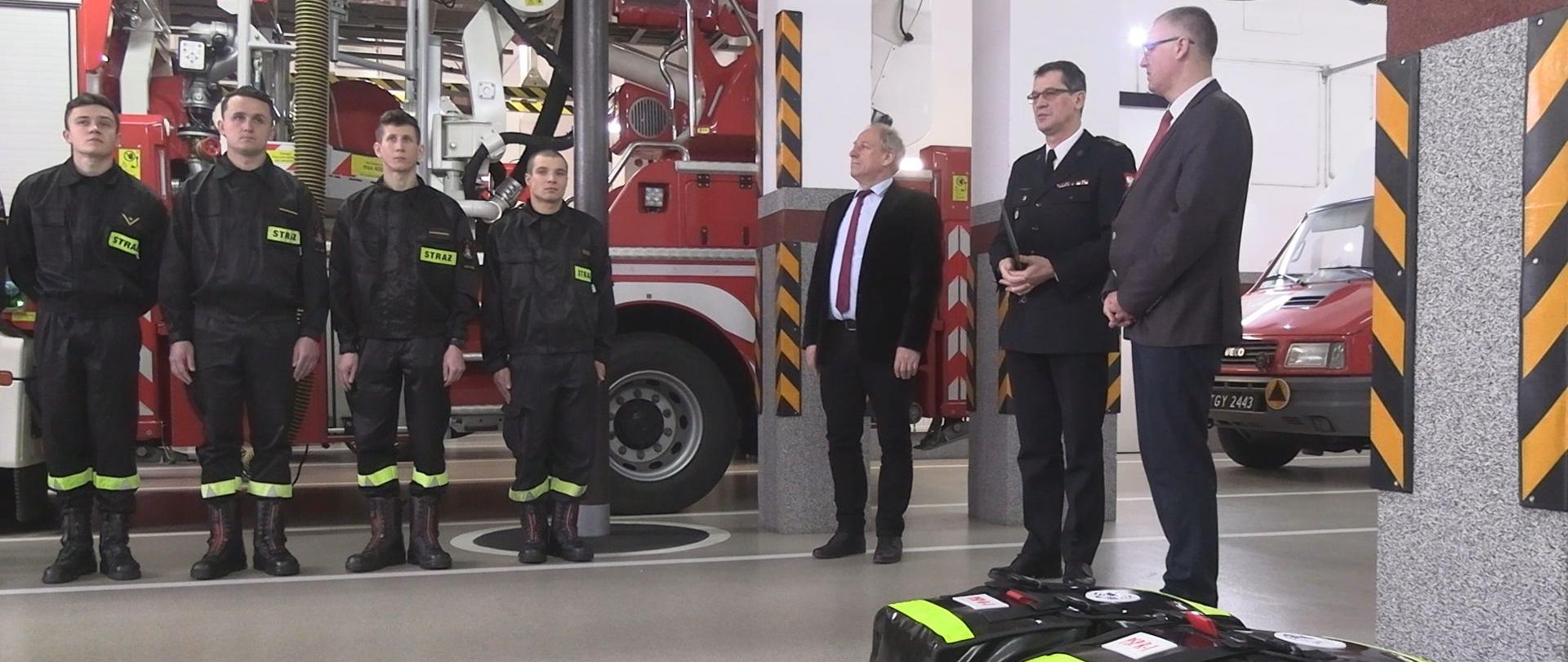 strażacy na garażu w remizie ustawieni w szeregu w postawie zasadniczej, obok nich stoi strażak w mundurze wyjściowym wraz z dwoma mężczyznami. Przed nimi ułożone dwie torby koloru czerwono-czarnego z napisem PSP-R1. 