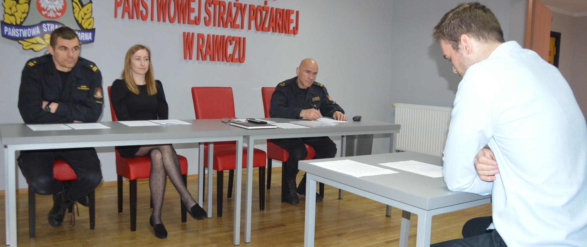 Nabór do służby. Świetlica Komendy Powiatowej PSP w Rawiczu. Komisja kwalifikacyjna przeprowadza czwarty etap naboru. Trzech członków komisji - dwóch umundurowanych i jedna osoba - cywil oceniają kandydata, który siedzi przy stoliku naprzeciwko komisji. Na ścianie napis Komenda Powiatowa PSP w Rawiczu i logo PSP.
