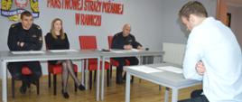 Nabór do służby. Świetlica Komendy Powiatowej PSP w Rawiczu. Komisja kwalifikacyjna przeprowadza czwarty etap naboru. Trzech członków komisji - dwóch umundurowanych i jedna osoba - cywil oceniają kandydata, który siedzi przy stoliku naprzeciwko komisji. Na ścianie napis Komenda Powiatowa PSP w Rawiczu i logo PSP.